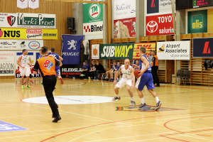 Ein Basketballfeld mit Spielern im Vordergrund und einer holzgetäfelten Wand mit Postern und Werbetafeln im Hintergrund.