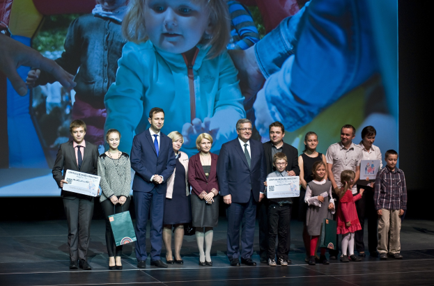 Gruppe von Menschen auf einer Bühne mit Taschen und Urkunden, mit einem Bildschirm im Hintergrund, der ein Kind und andere zeigt.