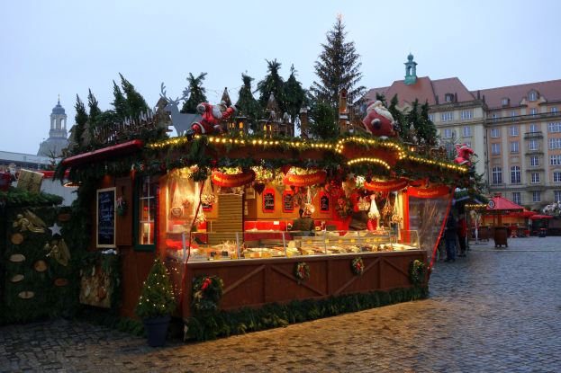 Ein gemütlicher Weihnachtsmarktstand auf einem Kopfsteinpflasterweg, umgeben von geschmückten Gebäuden, beleuchteten Bäumen und Menschen, mit dem Himmel im Hintergrund.