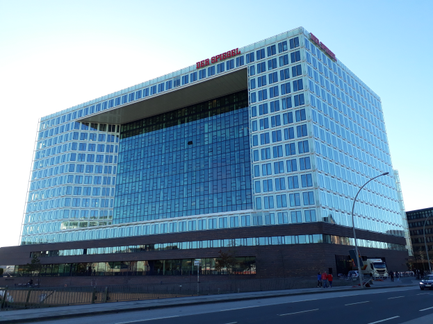 Ein großes Gebäude mit vielen Fenstern, das als Hauptsitz der Deutschen Bank identifiziert wurde, umgeben von Straßenmasten, Straßenlaternen, Kraftfahrzeugen, Füßgängern, Bäumen und einem bewölktem Himmel.