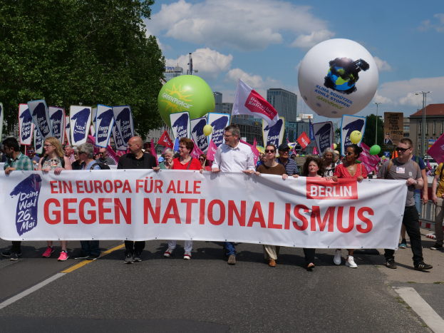 Eine Gruppe von Menschen marschiert auf einer Straße in Berlin, hält eine Fahne, Flaggen und Luftballons, mit Bäumen und Gebäuden auf der linken Seite und einem bewölkten Himmel im Hintergrund, nimmt an einer Demonstration gegen Nationalismus teil.