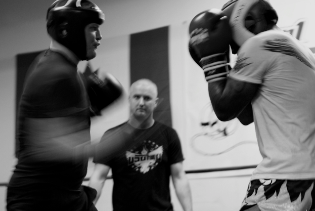 Zwei Boxer in Helmen und Handschuhen kämpfen im Ring, ein Schiedsrichter steht zwischen ihnen und ein Banner hängt an der Wand im Hintergrund.