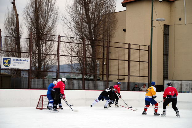 Menschen beim Eishockeyspielen auf einem Eisstadion mit Gebäuden, Bäumen, einer Straßenlaterne, einem Namensschild und Zäunen im Hintergrund unter einem Himmel.