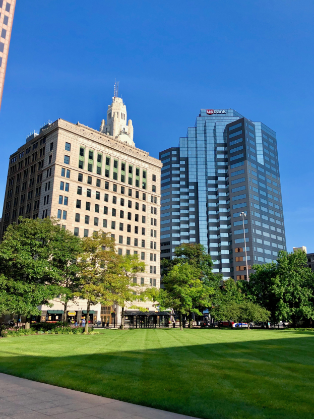 Ein Stadtpark mit Hochhäusern im Hintergrund, umgeben von grünem Gras, Bäumen und Laternenmasten, mit Menschen, die herumlaufen und dem Himmel darüber.