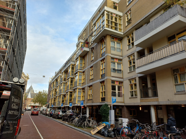 Eine Straße in Amsterdam mit parkenden Fahrrädern, fahrenden Fahrzeugen, Gebäuden mit Fenstern, Schildern auf Pfählen, Bäumen und einem bewölkten Himmel.