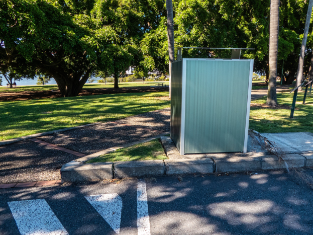 Transportable Toilette am Straßenrand in einem Park, umgeben von Bäumen und Gras, mit einem Wasserlauf und einem klaren blauen Himmel im Hintergrund.