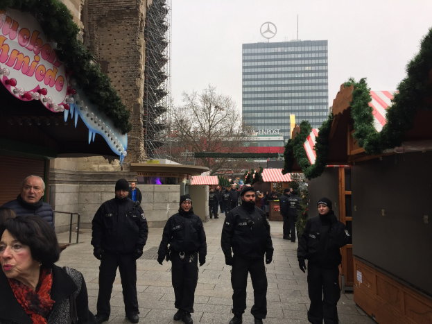 Polizeibeamte in schwarzen Uniformen stehen vor einem geschmützten Weihnachtsmarkt in Berlin, mit festlichen Dekorationen, Gebäuden, Bäumen und einem klaren blauen Himmel im Hintergrund.