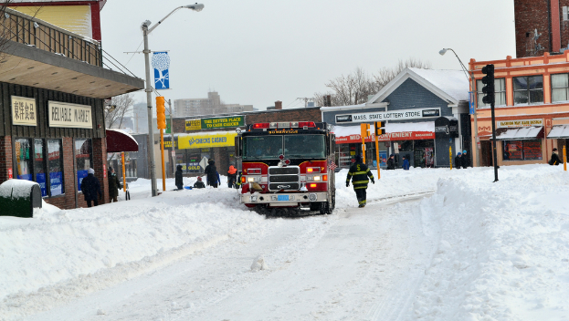 Eine Feuerwehrauto steht im Schnee vor Gebäuden, mit Geschäften und Laternenmasten links, einer Ampel in der Mitte und Schnee auf dem Boden.