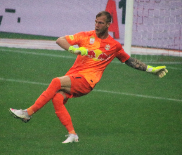 Ein professioneller Fußballspieler in einem orangen Trikot und Fußballhandschuhen kickt einen Fußball auf einem grünen Feld mit einem Netz und einer Texttafel im Hintergrund.