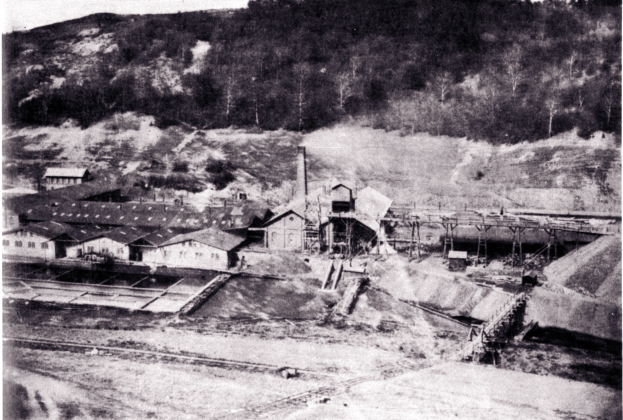 Schwarzes und wei├čes Foto eines Kohlebergbaus mit Geb├Ąuden, Strommasten und B├Ąumen im Hintergrund und Text unten.