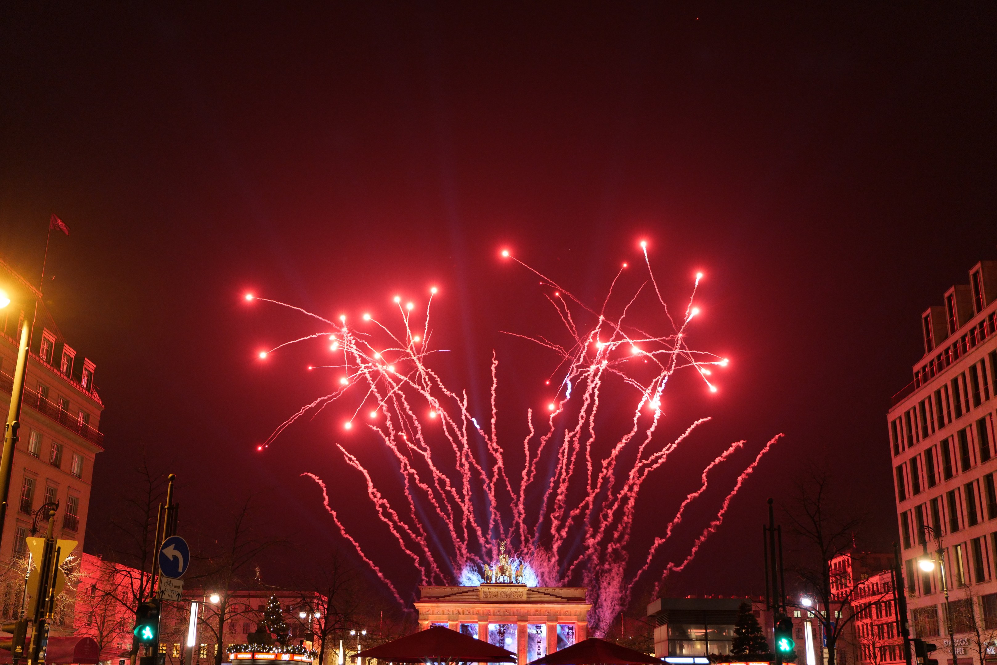 Eine belebte Stadtstraße an einem Neujahrsabend in Berlin, mit Gebäuden, Bäumen, Laternen, Verkehrszeichen, Zeltplanen, Menschen und einem prächtigen Feuerwerk am Himmel.