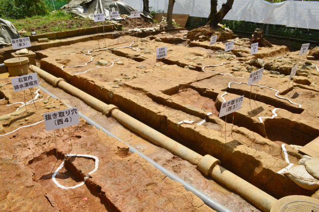 Große Grabungsstelle mit Erde, umgeben von Bäumen, Pflanzen, Gras, einem Zaun, einer Abdeckung und einer Wand, mit Schildern, Rohren und verstreuten Gegenständen.