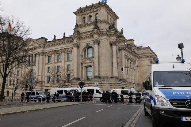 Eine Gruppe von Polizisten steht vor dem Reichstaggebäude in Berlin, Deutschland, mit Fahrzeugen, einem Zaun, Verkehrszeichen, Laternen, Bäumen und Flaggen im Hintergrund, bei klarem Himmel.