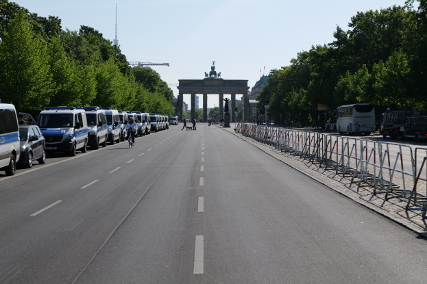 Eine lange Reihe von Polizeiwagen, die auf der Stra√üe vor dem Brandenburger Tor in Berlin, Deutschland, geparkt sind, mit Menschen, die Fahrr√§der fahren und auf der Stra√üe stehen, sowie Absperrungen und B√§ume, die die Seiten säumen, und ein Tor mit Statuen im Hintergrund.