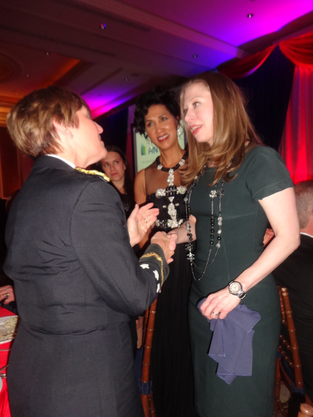 Vier Frauen, die sich im Vordergrund unterhalten, mit einem Stuhl rechts, einem Vorhang oben rechts, Lampen oben und einem roten Objekt links.