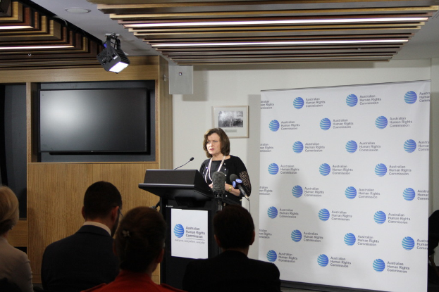 Eine Frau steht auf einer Bühne und spricht in ein Mikrofon, vor ihr sitzen Zuschauer, an der Wand hängt ein Banner, an der Wand steht ein Fernseher und an der Decke sind Lampen.