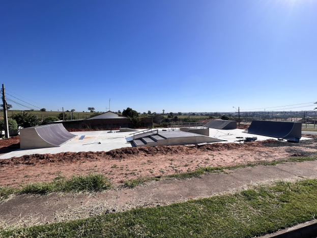 Skatepark mit mehreren Rampen, Grasfläche, Bäumen, Pfählen, Drähten und einem klaren blauen Himmel.