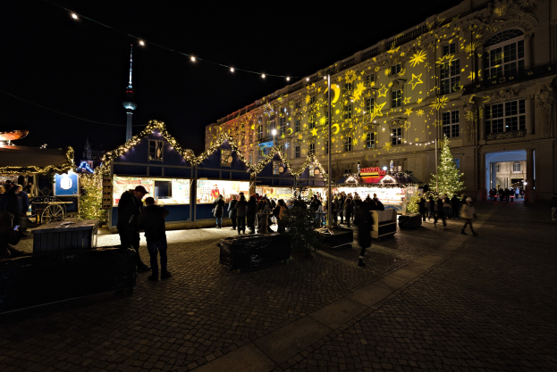 Ein geschäftiger Weihnachtsmarkt in Berlin, Deutschland, mit Menschen um dekorierte Stände und festlich geschmückte Gebäude im Hintergrund unter einem dunklen Himmel.