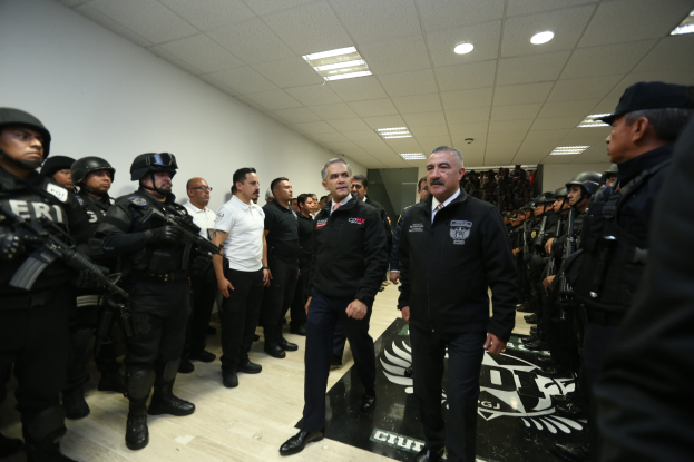 Eine Gruppe von Männern steht vor einer Reihe von bewaffneten, helmträgenden Polizeibeamten in einem Gerichtssaal, mit einer Wand und Deckenbeleuchtung im Hintergrund.