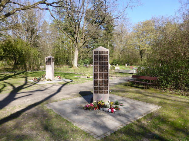 Kriegerdenkmal auf einer grünen Wiese mit Bäumen, umgeben von Blumensträußen und einer Bank, Gräber im Hintergrund unter einem klaren blauen Himmel.