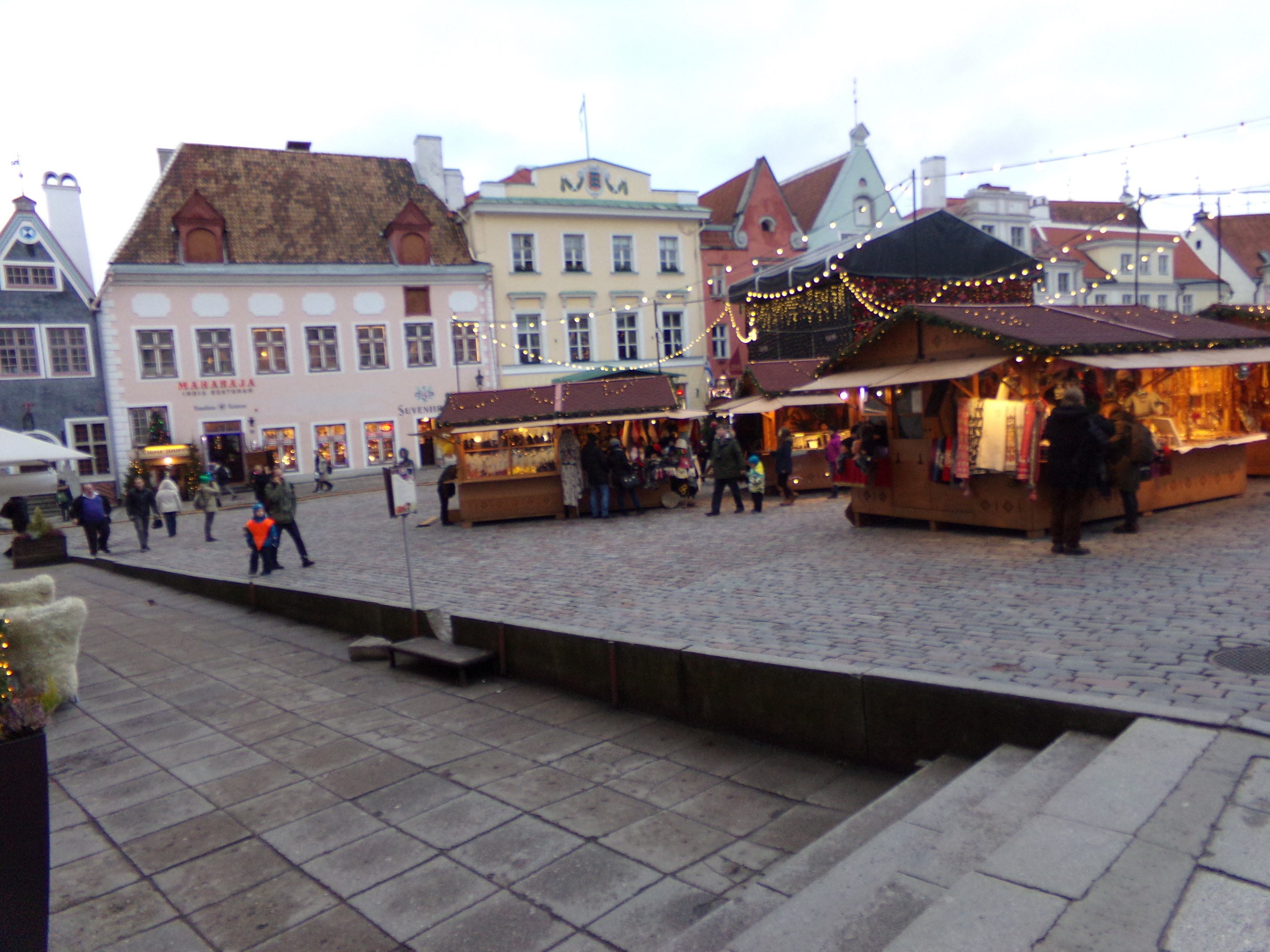 Ein geschäftiger Weihnachtsmarkt in Tallinn, Estland mit Menschen um geschmückte Stände, festliche Lichter, bewölkten Himmel, Stufen nach oben und Topfpflanzen im Bereich.
