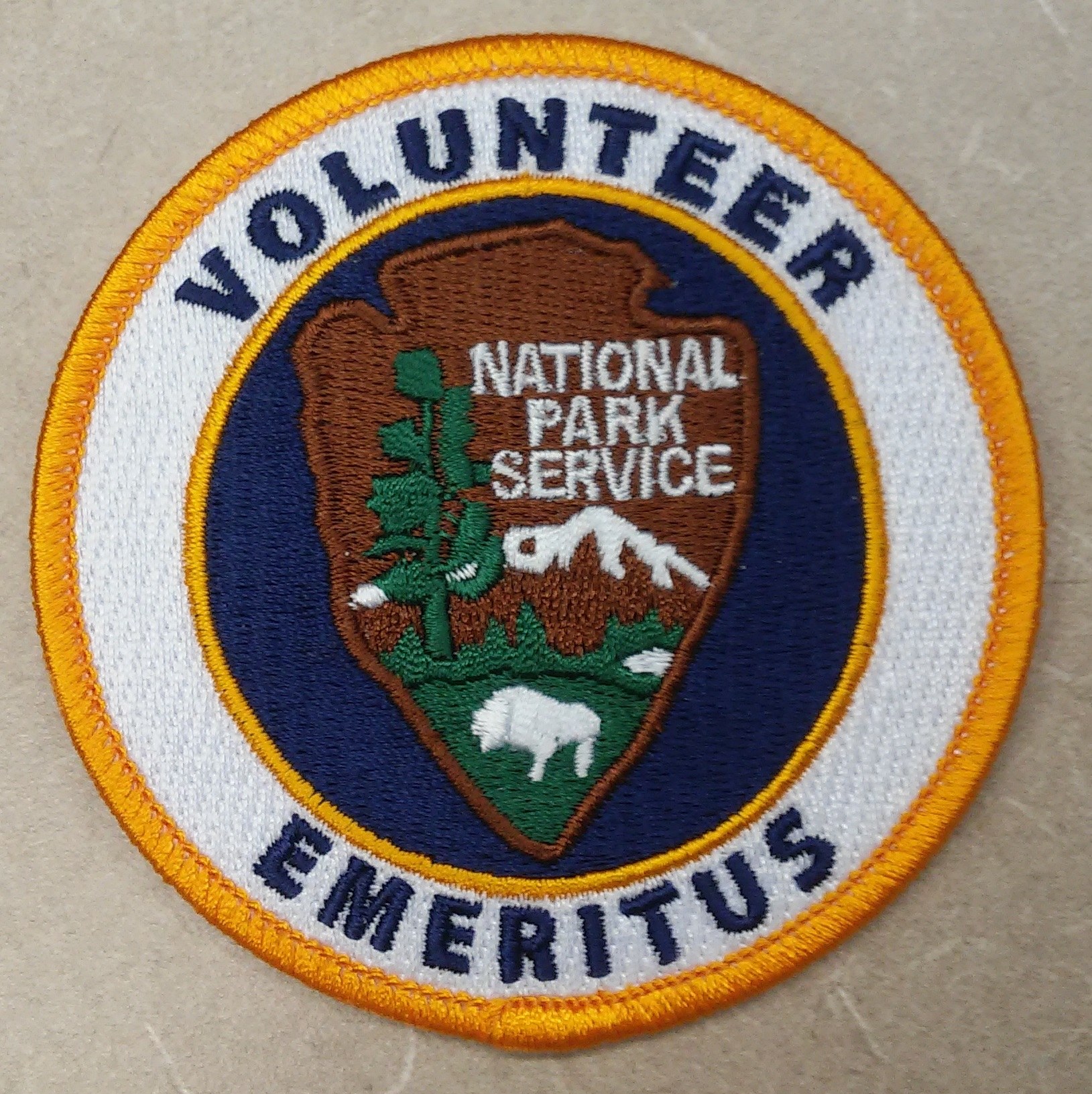 Runder Flicken mit blauem Rand und weißem Hintergrund, der mittig den schwarzen Text "Volunteer National Park Service Emeritus" in einem gelben Kreis enthält.