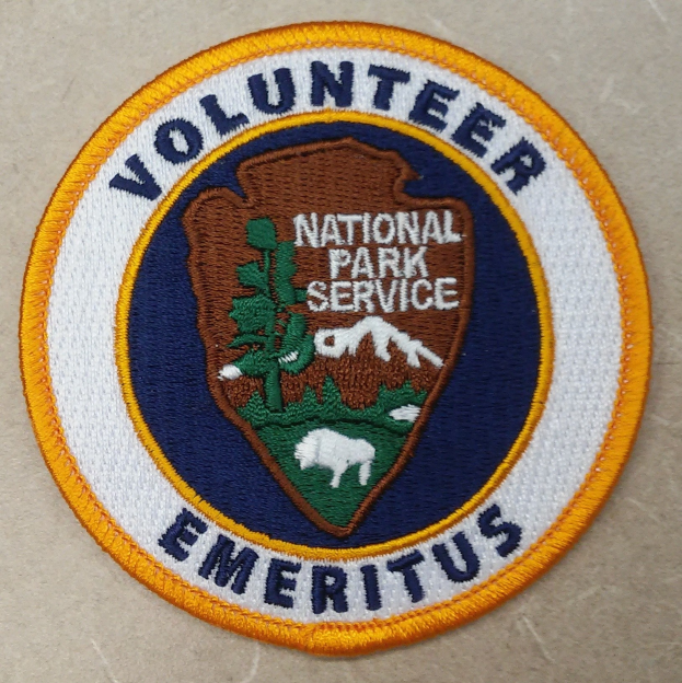 Runder Flicken mit blauem Rand und weißem Hintergrund, der mittig den schwarzen Text "Volunteer National Park Service Emeritus" in einem gelben Kreis enthält.