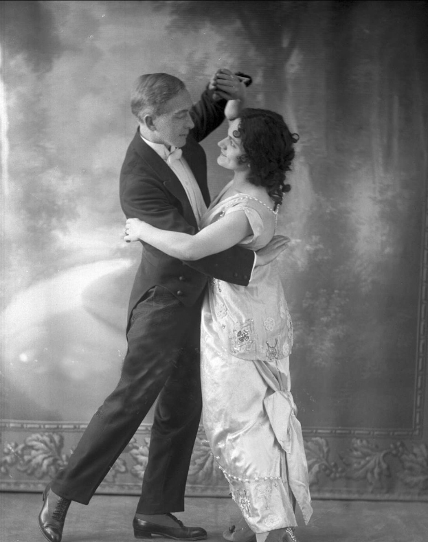Altes Schwarz-Weiß-Foto eines Mannes in einem Anzug und einer Frau, die Tango tanzen, mit kunstvollen Wanddesigns im Hintergrund.
