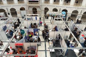 Eine große Gruppe von Menschen versammelt sich in einem Raum voller Stände, Banner, Tische, Stühle und Ballons, mit einem Gebäude mit Bögen, Säulen, Geländern und einem Bildschirm im Hintergrund.