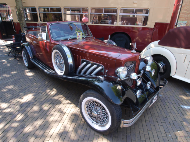 Ein Oldtimer neben einem Doppeldeckerbus auf einem Kopfsteinpflasterweg, mit Stühlen und einem Baum im Vordergrund, einem Gebäude im Hintergrund und Menschen im Bus.