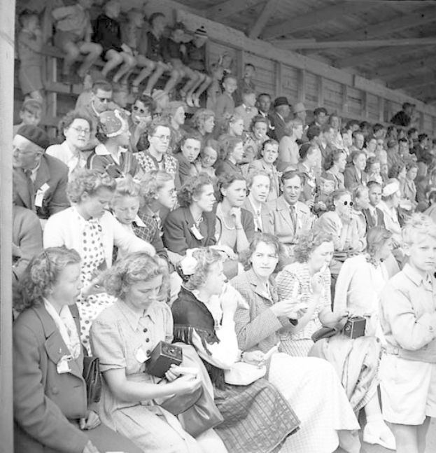 Schwarzes und weißes Foto einer Menge, die in einem Stadion mit einer hölzernen Wand und einem Dach sitzt, wobei einige Kameras halten.