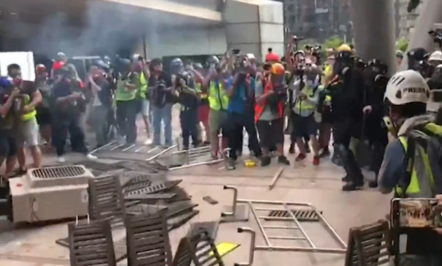 Menschen in Helmen und Jacken stehen vor einem raucherfüllten Gebäude, einige halten Kameras, mit verstreuten Metallobjekten und Stadtgebäuden im Hintergrund.