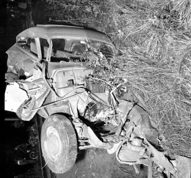 Ein Schwarz-Weiß-Foto eines schwer beschädigten Autos am Straßenrand, mit einer eingedrückten Front, zersplitterten Fenstern, einem Baum im Hintergrund und verstreuten Gegenständen am Tatort.