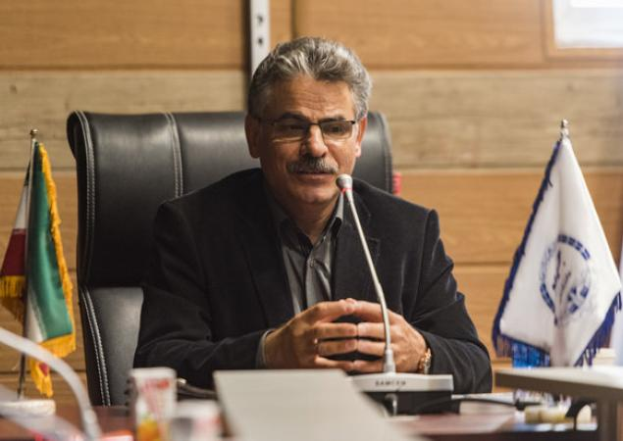 Ein Mann in einem schwarzen Blazer und Brille sitzt an einem Tisch mit einem Mikrofon, mehreren Flaggen und einer hölzernen Wand mit einem Fenster im Hintergrund.