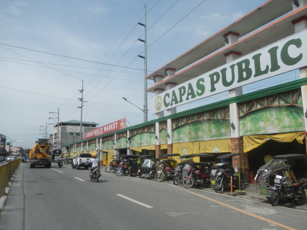 Eine belebte Stadtstraße mit Fahrzeugen, einem Geländer auf der linken Seite, Gebäuden, Pfählen, Laternen, Schildern und einigen Menschen, unter einem bewölkten Himmel, mit einem Gebäude mit der Aufschrift "Capas Public".
