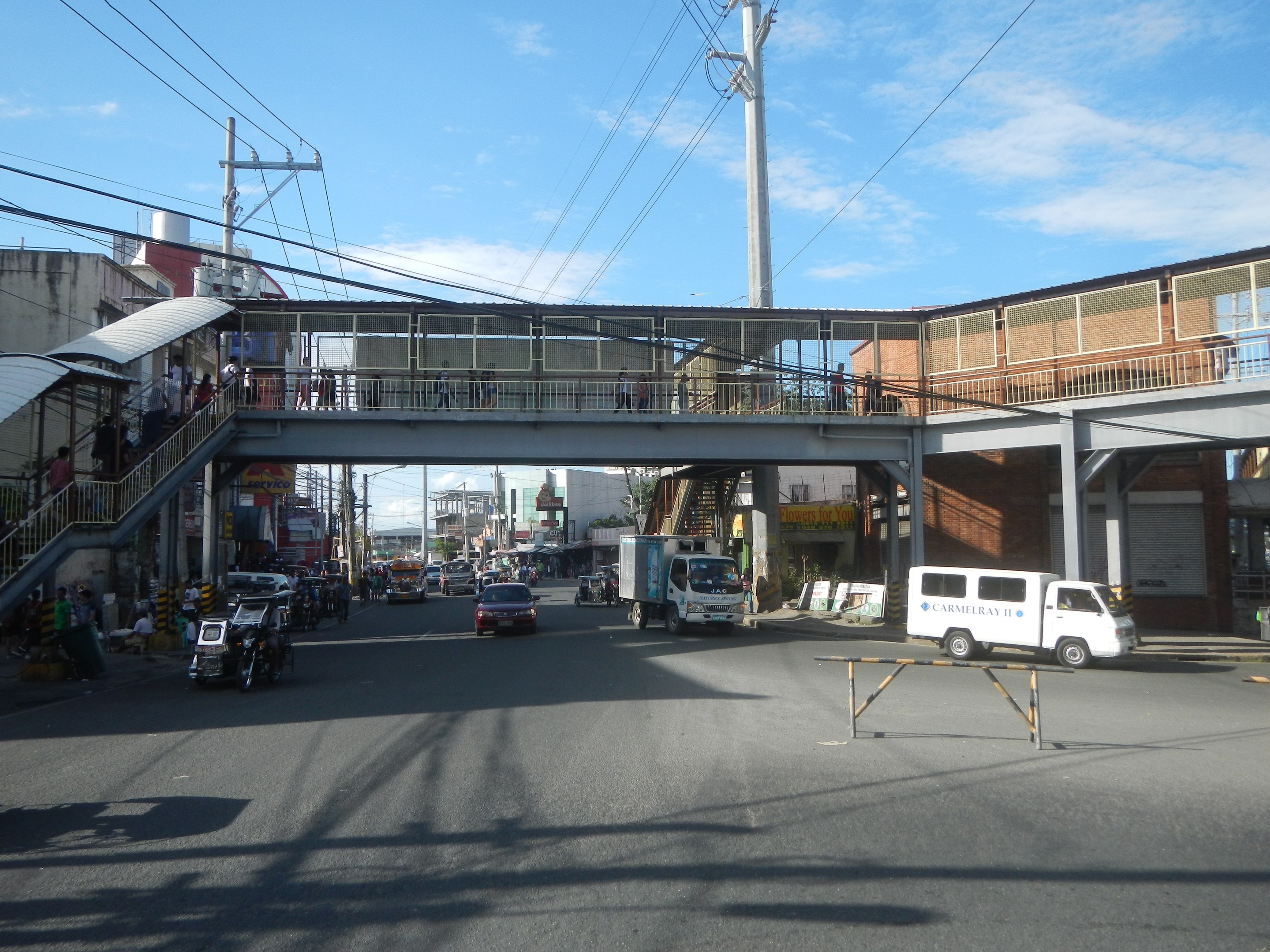 Stadtstraße mit Fahrzeugen, eine Fußgängerbrücke mit Menschen, Strommasten mit Drähten, Gebäude und ein bewölkter Himmel.