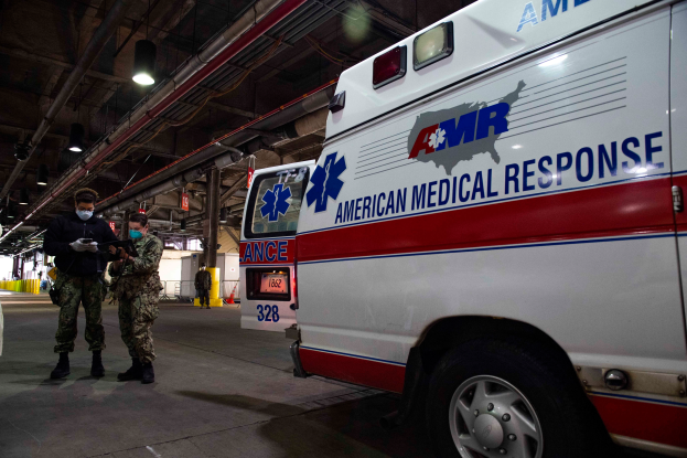 Ein Ambulanzfahrzeug der American Medical Response (AMR) steht in einem Gebäude mit zwei maskierten Personen davor, einem Fahrzeug auf der linken Seite und mehreren Menschen, Lampen und Rohren im Hintergrund.