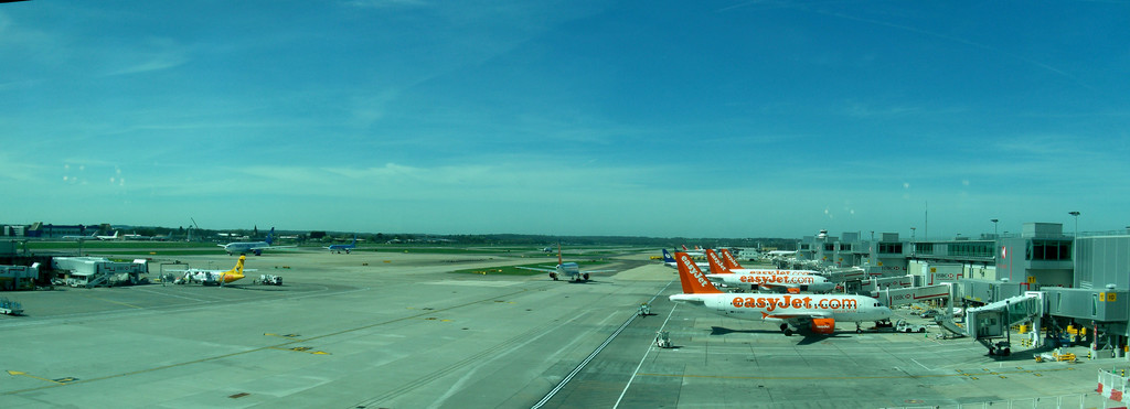 Eine Flughafen-Szene mit Flugzeugen, Gebäuden, Polen, Fahrzeugen, Bäumen und einem Himmel darüber sowie einer Straße darunter und einem Zaun im Vordergrund.