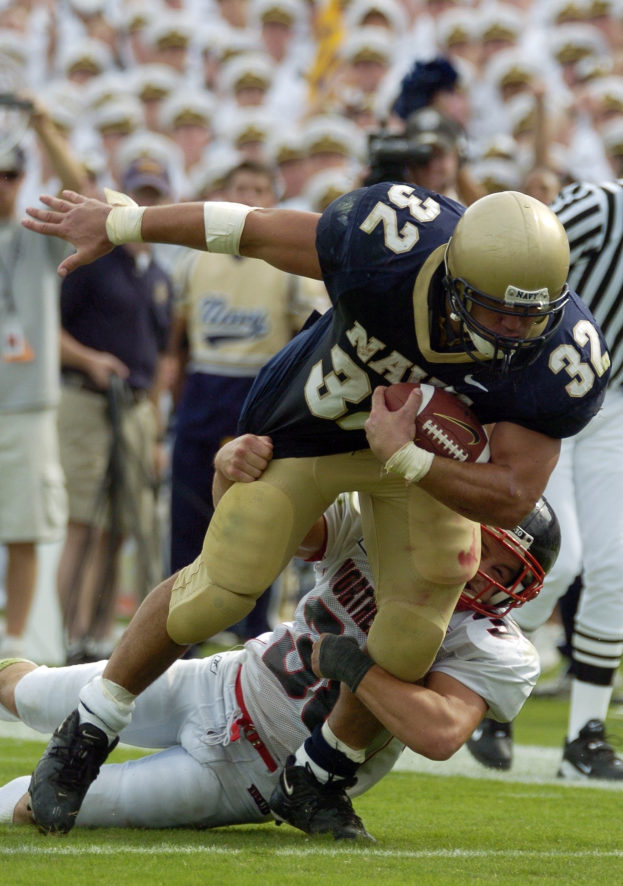 Zwei Fussballspieler auf dem Feld, einer in einem Sportsdress und Helm, der von einem anderen Spieler zu Fall gebracht wird, der am Boden liegt, mit anderen Spielern und Zuschauern im Hintergrund.