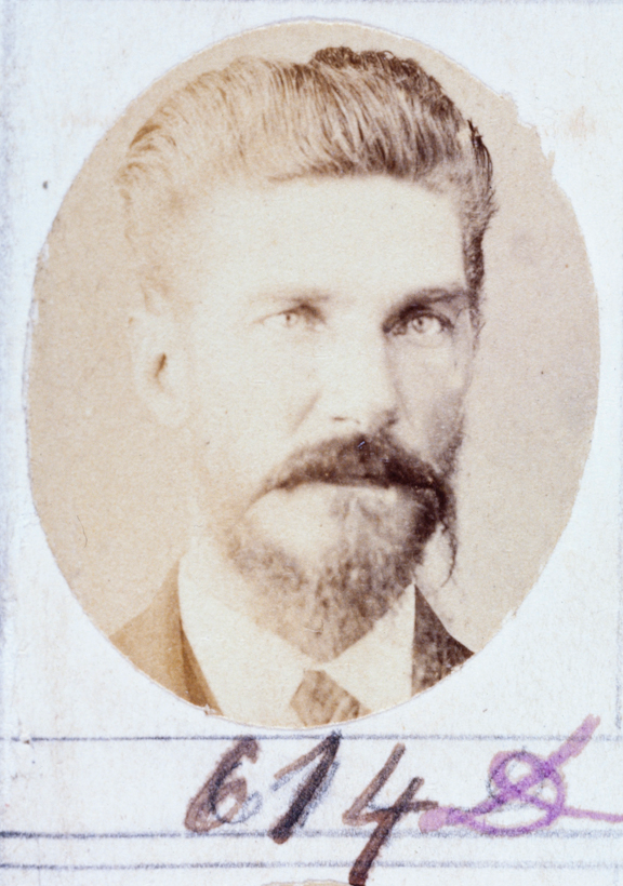 Ein altes Foto eines Mannes mit Bart und Schnurrbart, der direkt in die Kamera schaut und dessen Lippen leicht geöffnet sind, mit ordentlich gekämmtem Haar und einem gut gepflegten Bart.