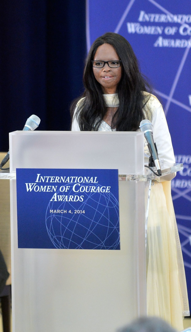 Frau in weißem Kleid und Brille, die vor einem Publikum an einem Podium mit zwei Mikrofonen und einer Tafel spricht, vor einem blauen Banner mit der Aufschrift "International Women of Courage Awards 2014" im Hintergrund.