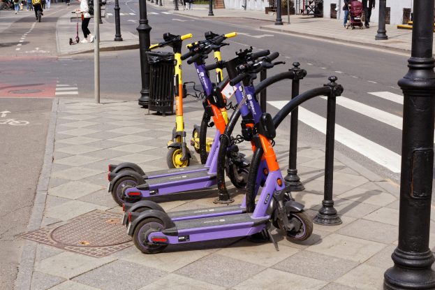 Eine Reihe von Elektro-Scootern, die an einer Stra├če geparkt sind, mit Passanten, Radfahrern, einem Müllcontainer im Vordergrund und Geb├Ąuden, B├Ąumen, Pflanzen und anderen Gegenst├Ąnden im Hintergrund.