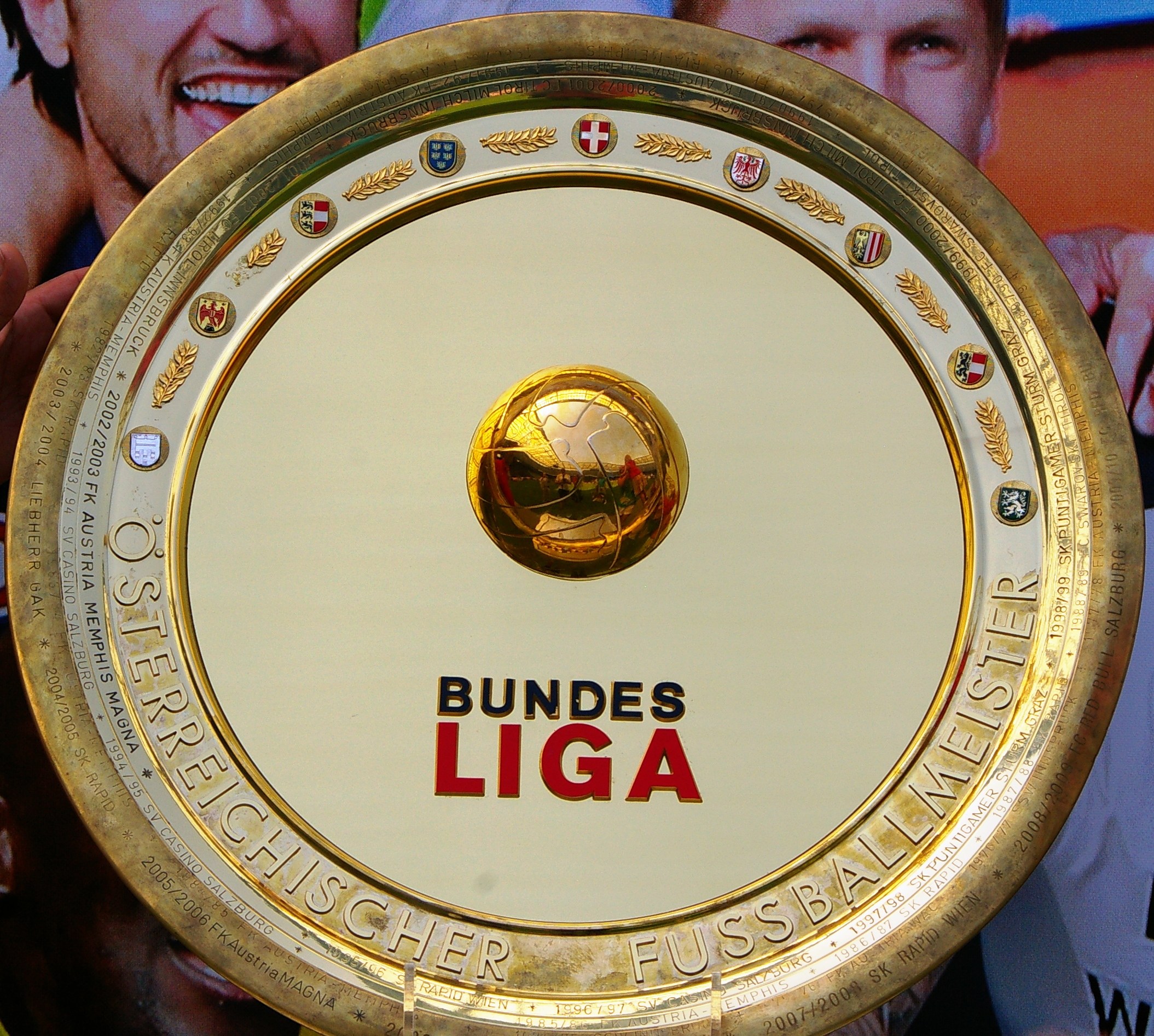 Goldener Schild mit "Bundesliga"-Text, gehalten von einem Mann mit einem Banner im Hintergrund mit drei Personen.