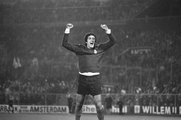 Ein Schwarz-Weiß-Foto eines Mannes, der auf einem Fußballfeld ein Tor feiert, mit erhobenen Armen, umgeben von Menschen, einem Zaun und Bannern mit Text.