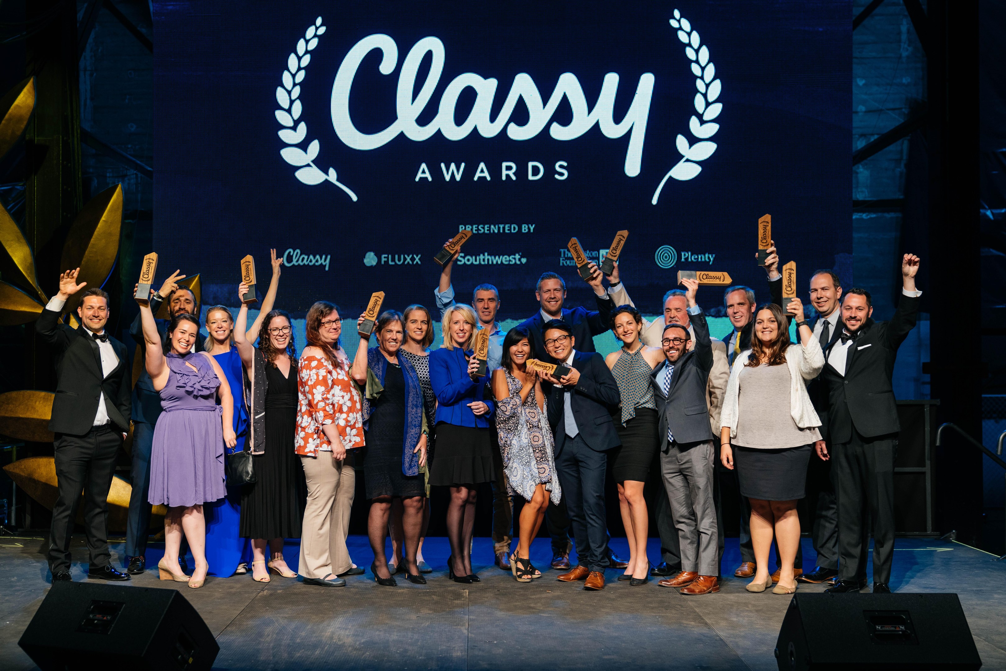 Gruppe von Menschen auf einer Bühne, die Auszeichnungen halten, mit Rednern darunter und einem Bildschirm im Hintergrund mit der Aufschrift "Classy Awards 2019".