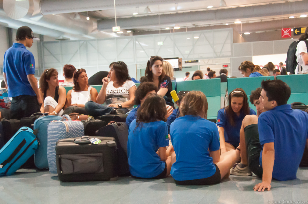 Ein belebtes Flughafeninnere mit vielen Menschen, die auf Flüge warten, Gepäck links und Glastüren mit Hinweisen im Hintergrund.