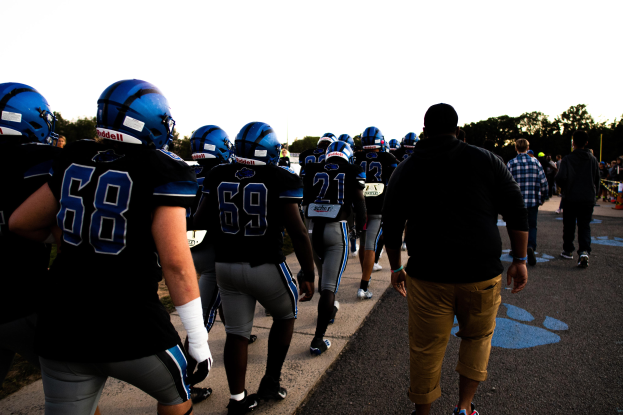 Eine Gruppe von Fussballspielern in Helmen und Stiefeln geht eine Strasse entlang mit Bäumen und einem klaren blauen Himmel im Hintergrund.