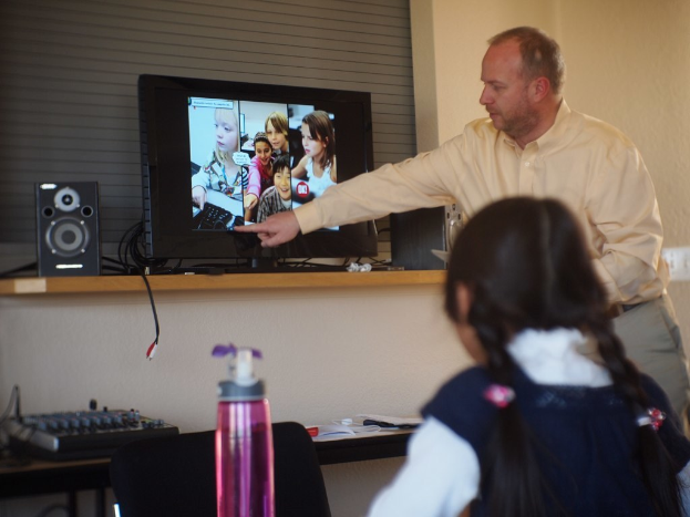 Ein Mann und ein Kind befinden sich in einem Raum mit einem Fernseher, einem Lautsprecher und einer Musikanlage; der Mann erklärt etwas aus dem Fernseher, und es gibt eine Flasche links neben dem Kind.