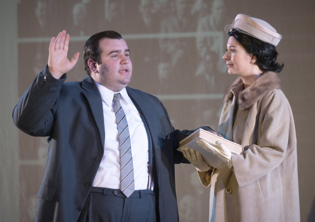 Ein Mann im Anzug und Krawatte und eine Frau mit einem Buch stehen zusammen auf einer Bühne mit einem Bildschirm im Hintergrund.