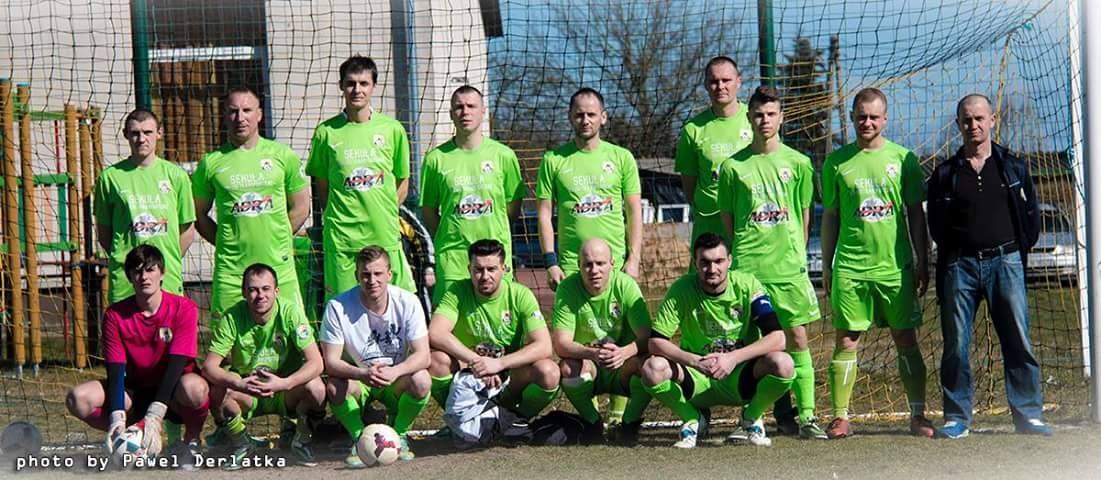 Gruppe von Männern in grünen T-Shirts auf einem Fußballfeld neben einem Torpfosten, mit einem Fußball auf dem Boden.